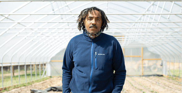 A man stands inside a greenhouse, looking directly at the camera without smiling.