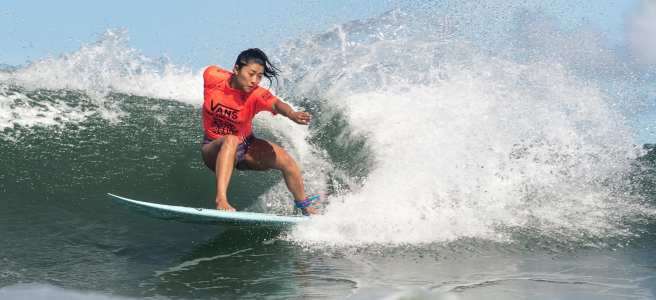 A surfer wearing a red "Vans" shirt rides a wave.