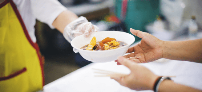 A chef wearing a yellow apron and a plastic glove hands a bowl of food to a customer who holds a pair of chopsticks.