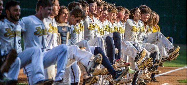 About twenty uniformed Savannah Bananas players line up side-by-side on the field doing a kick-line dance.
