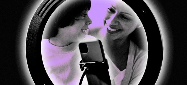 A small child sits on the lap of a woman; they gaze at one another, smiling. In front of them, a ring light and a smartphone on a gooseneck stand.