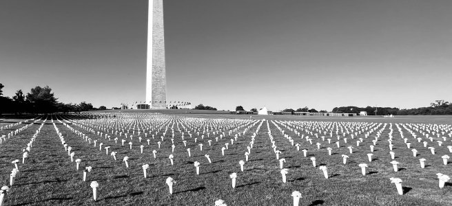 A flat, grassy field lined with rows of simple grave markers that form a geometric pattern; the Washington Monument stands in the background.