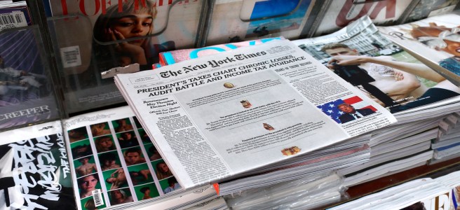 Stacks of thick and glossy fashion magazines on a newstand. A copy of the New York Times is displayed on top of the magazine stacks.