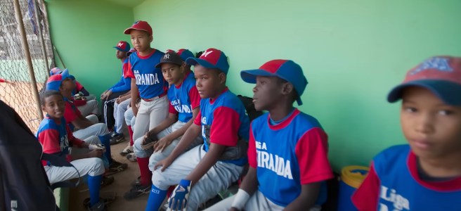 Nine Dominican youth league players, in uniform, sitting in their dugout.