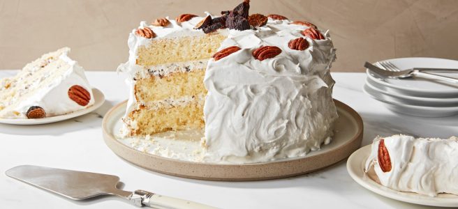 In the center of a white table is a 3-layer yellow cake on a rimmed plate, with thick white frosting spread with peaks. The top of the cake is ringed with pecan halves, and several ripe figs adorn the center. Two slices cut from the cake are on plates on the table, along with a few more plates, forks, and a cake server.